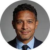 Smiling man with short curly hair wearing a dark suit and blue shirt against a gray background.